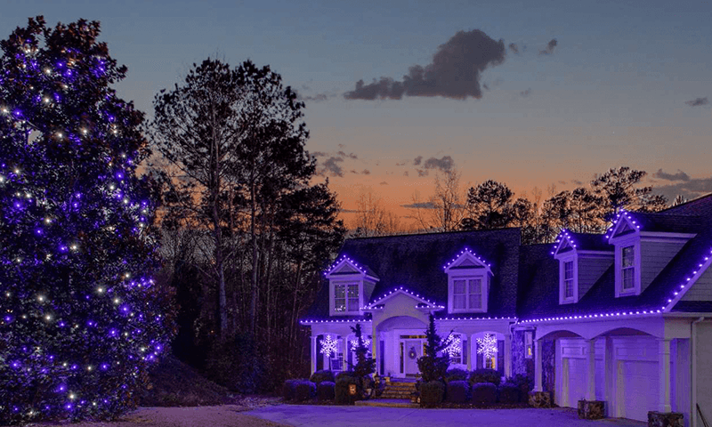 Home decorated with purple and white Christmas lights outlining the roofline and porch. A large front yard tree glows with matching purple and white lights, while oversized snowflake decorations add a festive accent. | Icy Lights LLC